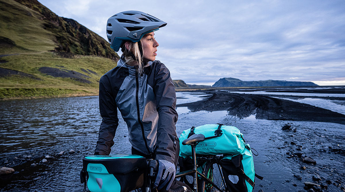 Radfahrerin mit ORTLIEB-Taschen, trägt einen Helm, steht an einem Fluss in einer zerklüfteten, bergigen Landschaft.