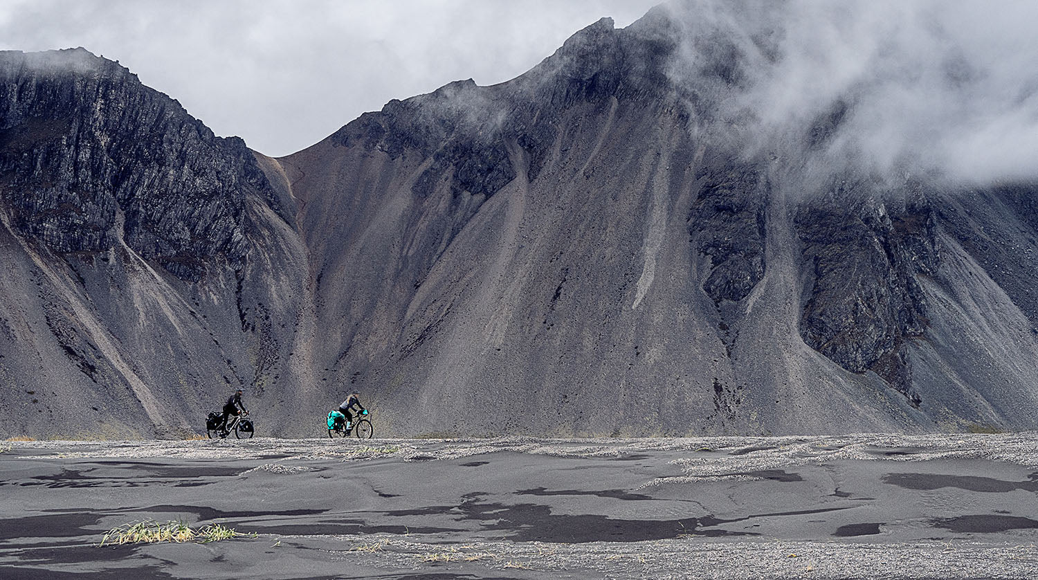 Zwei Radfahrer fahren durch eine karge Landschaft mit rauen, zerklüfteten Bergen und Nebel im Hintergrund.
