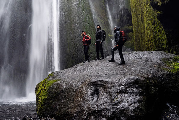 Drei Wanderer stehen auf einem nassen Felsen in der Nähe eines Wasserfalls, umgeben von moosbedeckten Klippen.
