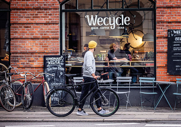 Eine Person mit einem Fahrrad steht vor Wecycle, einem Café und Fahrradgeschäft mit der Aufschrift „Bikes, Coffee, Gear“ und Menschen im Geschäft.
