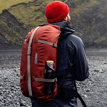 Person, die einen roten ORTLIEB Atrack Rucksack mit einer Wasserflasche in der Seitentasche trägt, steht im Freien in einer rauen, regnerischen Landschaft
