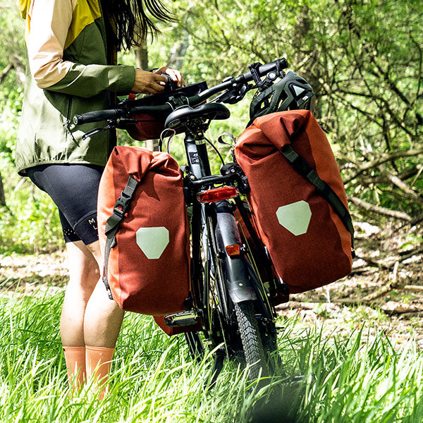 Person, die neben einem Fahrrad steht, das mit zwei großen roten ORTLIEB-Taschen auf der Vorderseite ausgestattet ist, in einem grasbewachsenen Waldgebiet.
