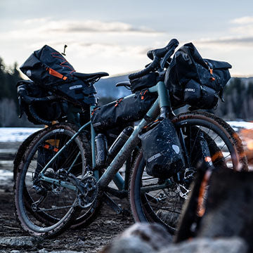 Zwei Fahrräder, ausgestattet mit ORTLIEB Bikepacking-Taschen, stehen im Freien auf einem schlammigen Weg in der Nähe eines Lagerfeuers, mit Bergen und Wald im Hintergrund.