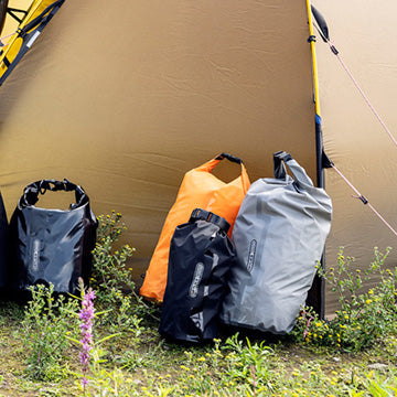 Mehrere ORTLIEB Packsäcke in verschiedenen Farben, darunter schwarz, orange und grau, vor einem Zelt auf einer Wiese auf dem Campingplatz