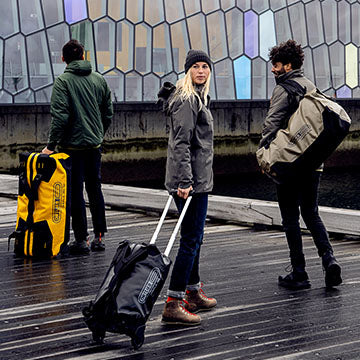 Drei Personen stehen im Freien mit ORTLIEB Duffles, eine trägt eine gelbe Duffle, während zwei andere schwarze und graue Duffles auf Rädern in der Nähe eines modernen Gebäudes ziehen.
